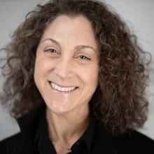 photo of a smiling middle aged white woman with brown shoulder length curly hair, wearing a black collared shirt